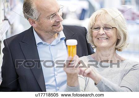 Stock Photography - Smiling senior woman showing pill bottle label to mature man in pharmacy.. Fotosearch