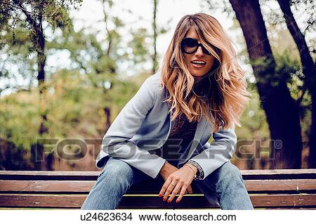 Stylish woman with long blond hair wearing sunglasses sitting on park bench, portrait View Large Photo Image Picture - Stylish woman with long blond hair wearing sunglasses sitting on park bench, portrait. Fotosearch