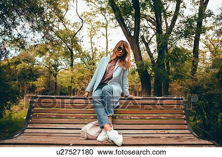 Stock Image - Stylish woman with long blond hair sitting on park bench, full length portrait. Fotosearch