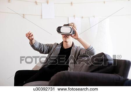 Stock Image - Teenage boy on sofa looking through virtual reality headset and reaching out. Fotosearch