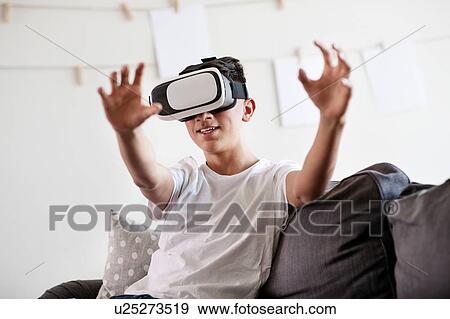 Stock Photo - Teenage boy on sofa looking through virtual reality headset and reaching out. Fotosearch
