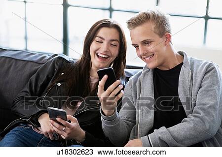 Stock Image - Teenage girl and boy sitting on sofa looking at smartphone. Fotosearch