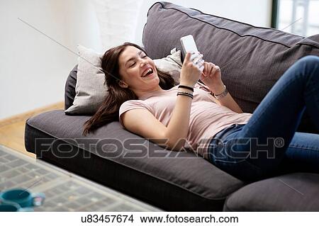 Picture - Teenage girl laughing on sofa looking at smartphone. Fotosearch