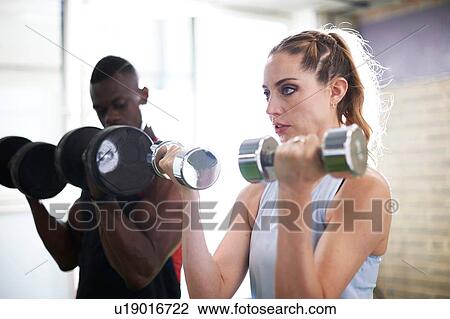 Stock Image - Trainer and female client lifting dumbbells in gym. Fotosearch