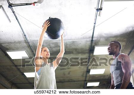 Stock Image - Trainer watching female client lift medicine ball in gym. Fotosearch