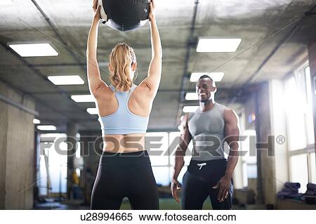 Stock Photography - Trainer watching female client lift medicine ball in gym. Fotosearch