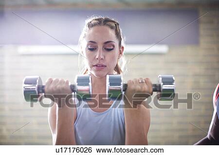 Stock Photograph - Trainer watching female client lifting dumbbells in gym. Fotosearch
