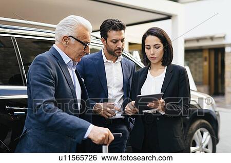 Stock Photograph - Two businessmen and woman outside hotel looking at digital tablet. Fotosearch