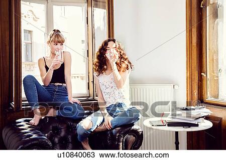Stock Image - Two young female friends sitting on living room armchair drinking water. Fotosearch