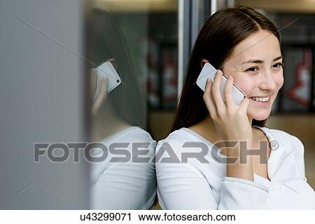 Stock Image - Woman leaning on glass wall using cellphone. Fotosearch