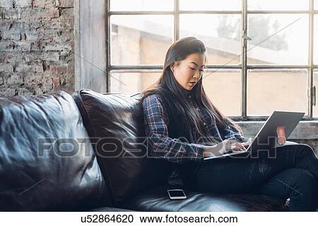 Stock Image - Woman on sofa by window using laptop. Fotosearch