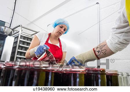 Stock Image - Woman pouring fresh jam into jars in industrial kitchen. Fotosearch