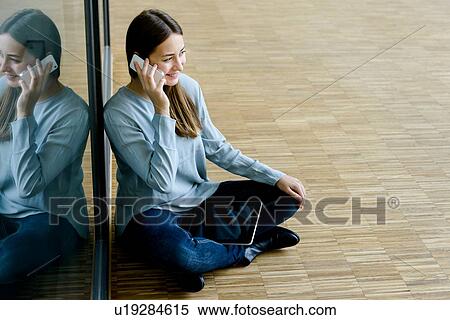 Stock Photography - Woman sitting cross legged by glass wall using cellphone. Fotosearch