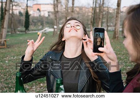 Stock Photo - Woman taking photograph of friend balancing bottle caps on cheeks. Fotosearch