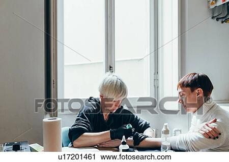 Stock Image - Woman tattooing friend's arm at home. Fotosearch