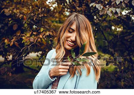 Stock Photo - Woman with long blond hair holding sprig of leaves in park, portrait. Fotosearch