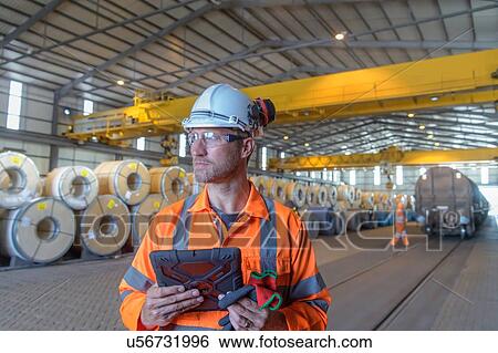 Stock Photograph - Worker using digital tablet by rows of sheet steel in storage at port. Fotosearch