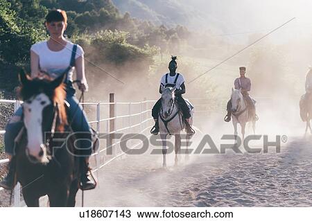 Stock Image - Young adults riding horses in dusty rural equestrian arena. Fotosearch
