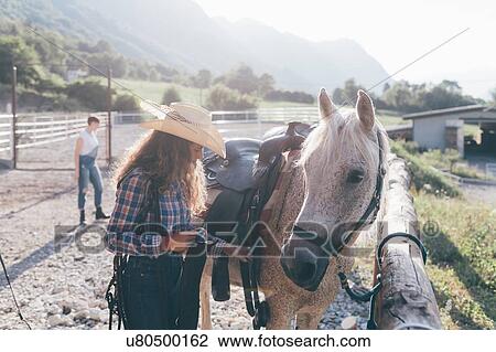 Stock Image - Young cowgirl checking saddle in rural equestrian arena. Fotosearch