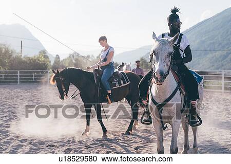 Stock Image - Young hipster man and woman riding horses in rural equestrian arena, Primaluna, Trentino-Alto Adige, Italy. Fotosearch
