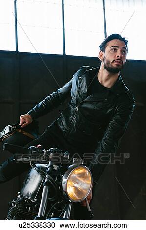 Stock Image - Young male motorcyclist mounting vintage motorcycle in garage. Fotosearch