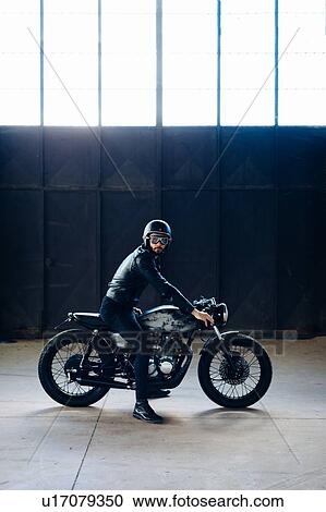 Stock Image - Young male motorcyclist on vintage motorcycle in empty warehouse, portrait. Fotosearch