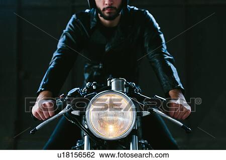 Stock Image - Young male motorcyclist straddling vintage motorcycle in garage, cropped front view. Fotosearch