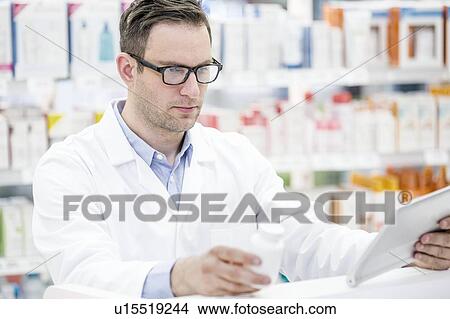 Young male pharmacist using digital tablet to check pill bottle in pharmacy. View Large Photo Image Picture - Young male pharmacist using digital tablet to check pill bottle in pharmacy.. Fotosearch