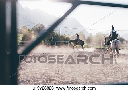 Stock Image - Young man and woman horse riding in rural equestrian arena. Fotosearch