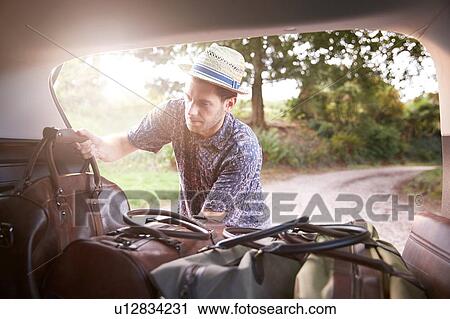 Stock Image - Young man in trilby removing luggage from car boot on rural road. Fotosearch