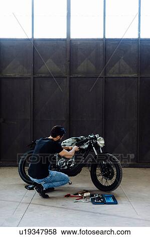 Young man kneeling to do maintenance on vintage motorcycle in empty warehouse View Large Photo Image Stock Photo - Young man kneeling to do maintenance on vintage motorcycle in empty warehouse. Fotosearch
