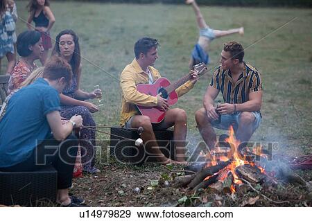 Stock Photo - Young man playing guitar at bonfire party in park. Fotosearch