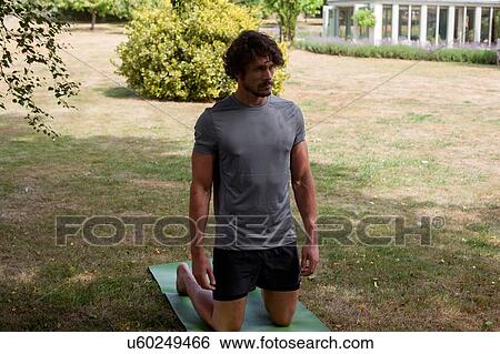 Stock Photograph - Young man practicing yoga in garden, kneeling on yoga mat. Fotosearch
