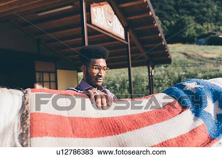 Stock Image - Young man putting american flag blanket on horse's back at ranch. Fotosearch