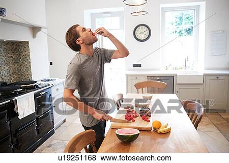 Stock Photo - Young man slicing and eating fresh fruit at kitchen table. Fotosearch