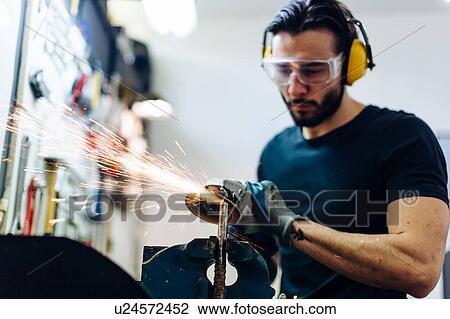 Stock Image - Young man using angle grinder on metal in workshop. Fotosearch