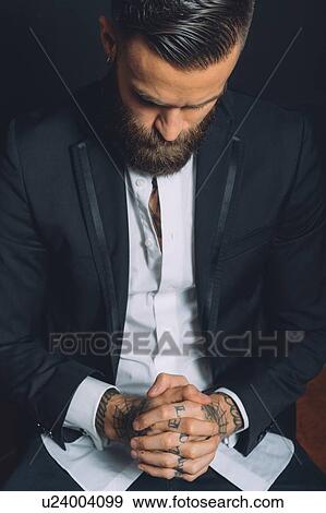Stock Photo - Young man wearing suit, sitting with hands clasped, tattoos on hands, pensive expression. Fotosearch
