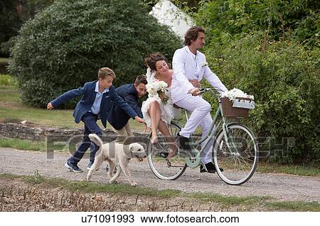 Stock Image - Young wedding guests running after newlyweds on bicycles. Fotosearch