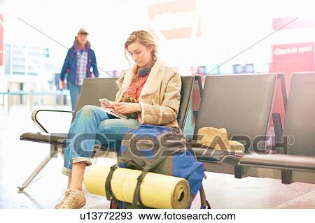 Stock Image - Young woman at airport, sitting with backpack beside her, using smartphone. Fotosearch