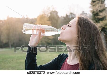 Stock Image - Young woman drinking from water bottle in park. Fotosearch