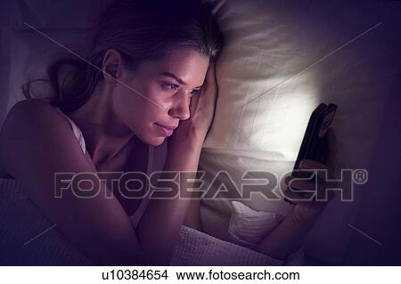 Picture - Young woman looking at smartphone in bed. Fotosearch