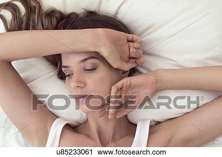 Stock Image - Young woman lying in bed with hand on head. Fotosearch