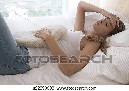 Stock Photo - Young woman on bed with hot water bottle and hand on head. Fotosearch