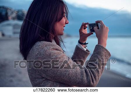 Stock Image - Young woman standing on beach, taking photograph of view, using smartphone. Fotosearch