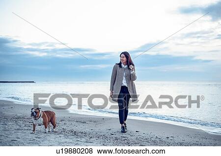 Stock Photo - Young woman walking along beach with pet dog. Fotosearch