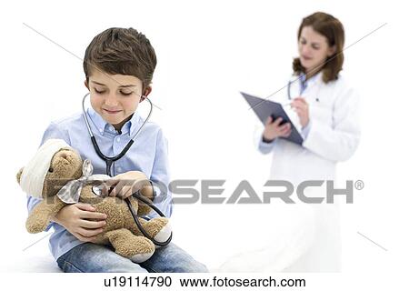 Stock Image - Boy role playing with teddy bear and stethoscope with doctor in background.. Fotosearch