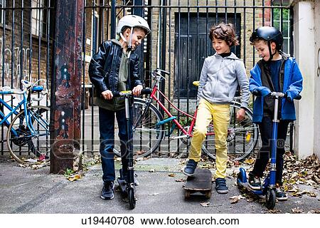 Stock Photo - Boys riding push scooter and skateboard, bicycles in background. Fotosearch