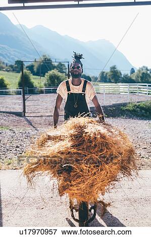Stock Photo - Cool young man with wheelbarrow hay in rural equestrian arena, portrait. Fotosearch