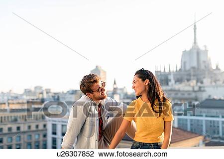 Stock Image - Couple enjoying sitting on edge of rooftop terrace, Milan, Italy. Fotosearch