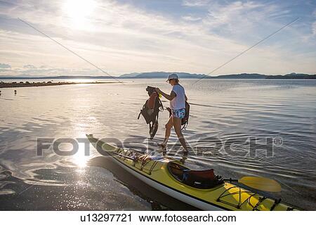 Stock Image - Mature female kayaker carrying life jackets from kayak, Quadra Island, Campbell River, Canada. Fotosearch